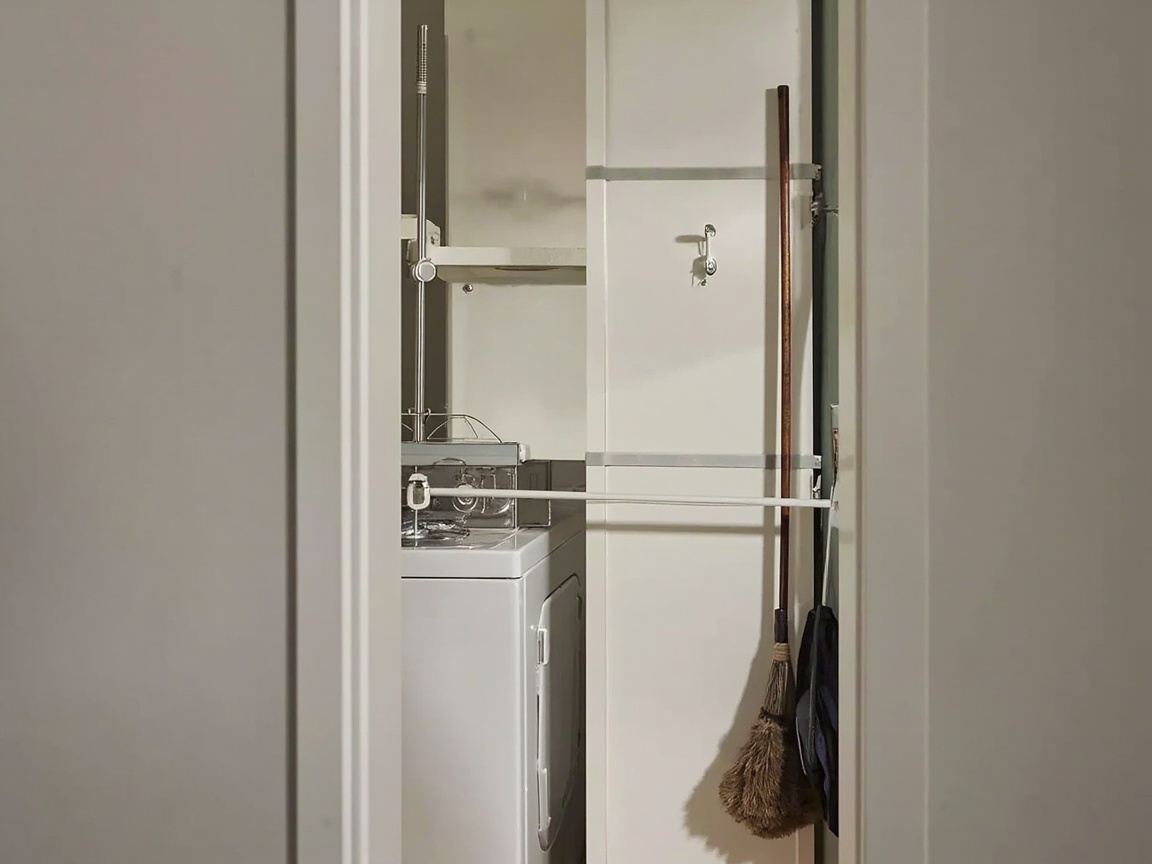 Vertical wall strip holding a broom, mop, duster, and a hanging iron behind a stacked washer and dryer